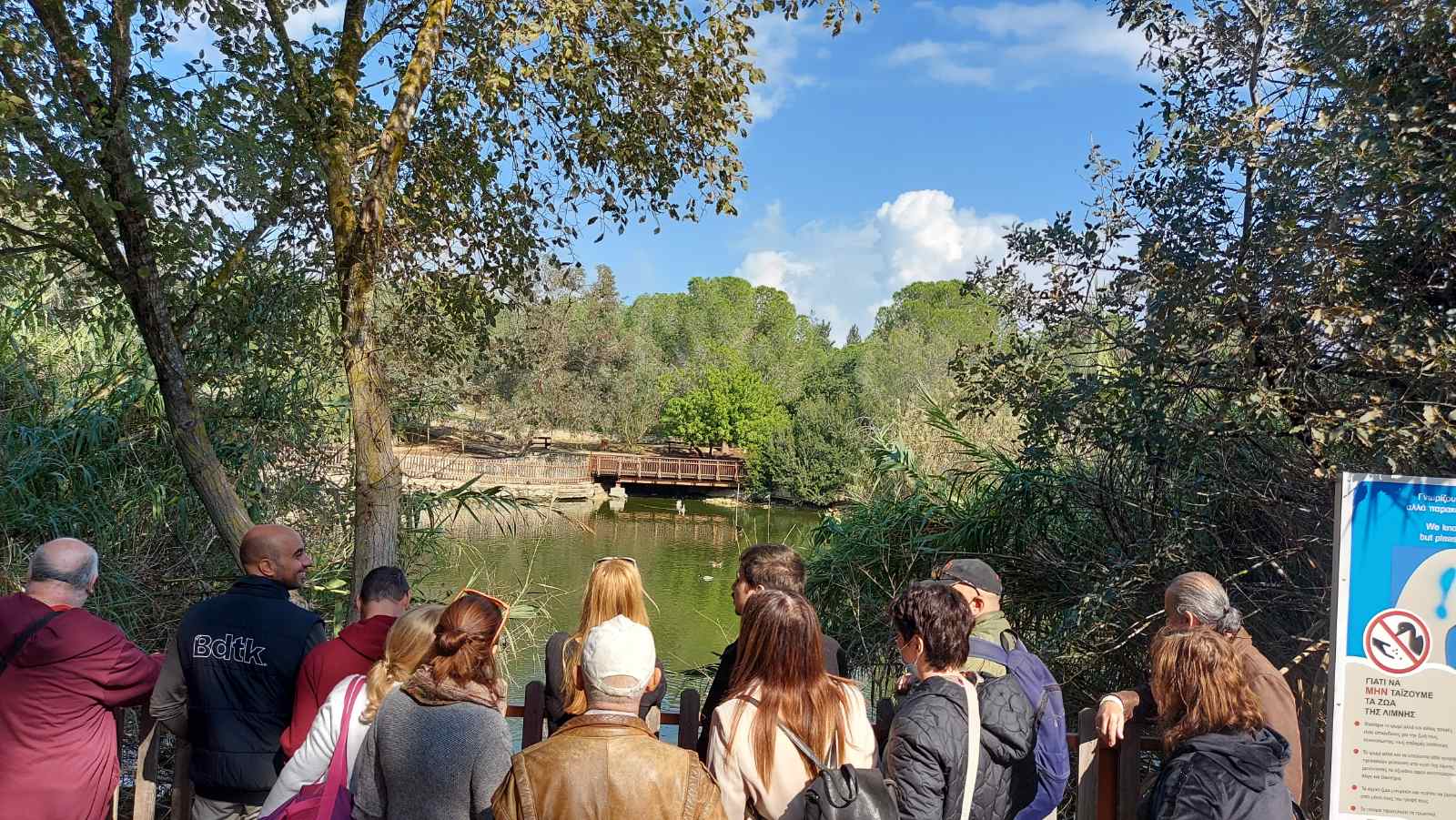 Οι εκπαιδευόμενοι είναι στο χώρο της λίμνης, που περιβάλλεται από ψηλά δένδρα. Πάπιες κολυμπούν στο νερό, ενώ στο βάθος, απέναντι από τους εκπαιδευόμενους βρίσκεται μια ξύλινη γέφυρα.